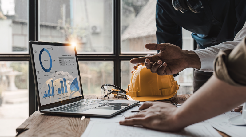 Top 10 Construction Project Management Tools for 2024: Streamline Your Builds Image of two men stood at a desk with a laptop on displaying different graphs on the screen to support construction project management tools article